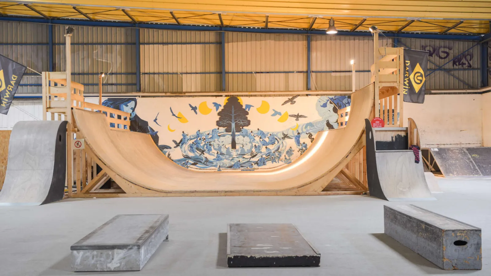 Indoor skate park at Bordeaux Darwin featuring a large wooden ramp with blue and yellow decorative motifs, wooden walkways, and ground-level skate obstacles.