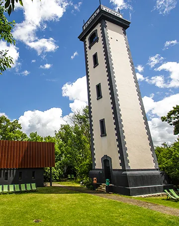 Tall white and black tower on Patiras Island in Bordeaux, surrounded by green lawn and a red building in the background, under a partly cloudy blue sky.