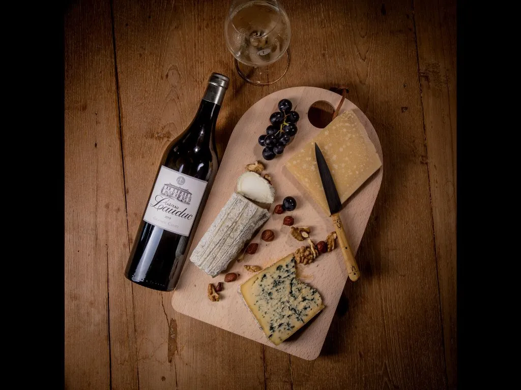 Wine and cheese tasting board featuring a bottle of Château Lauduc red wine, assorted cheeses including blue cheese, black grapes, nuts, bread and a wine glass on a light wooden board, set on dark wooden table.