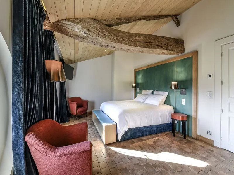 Elegant bedroom at Château Beauregard in Bordeaux featuring exposed wooden beams, white bed with green headboard, red armchairs, black curtains, and tiled flooring.