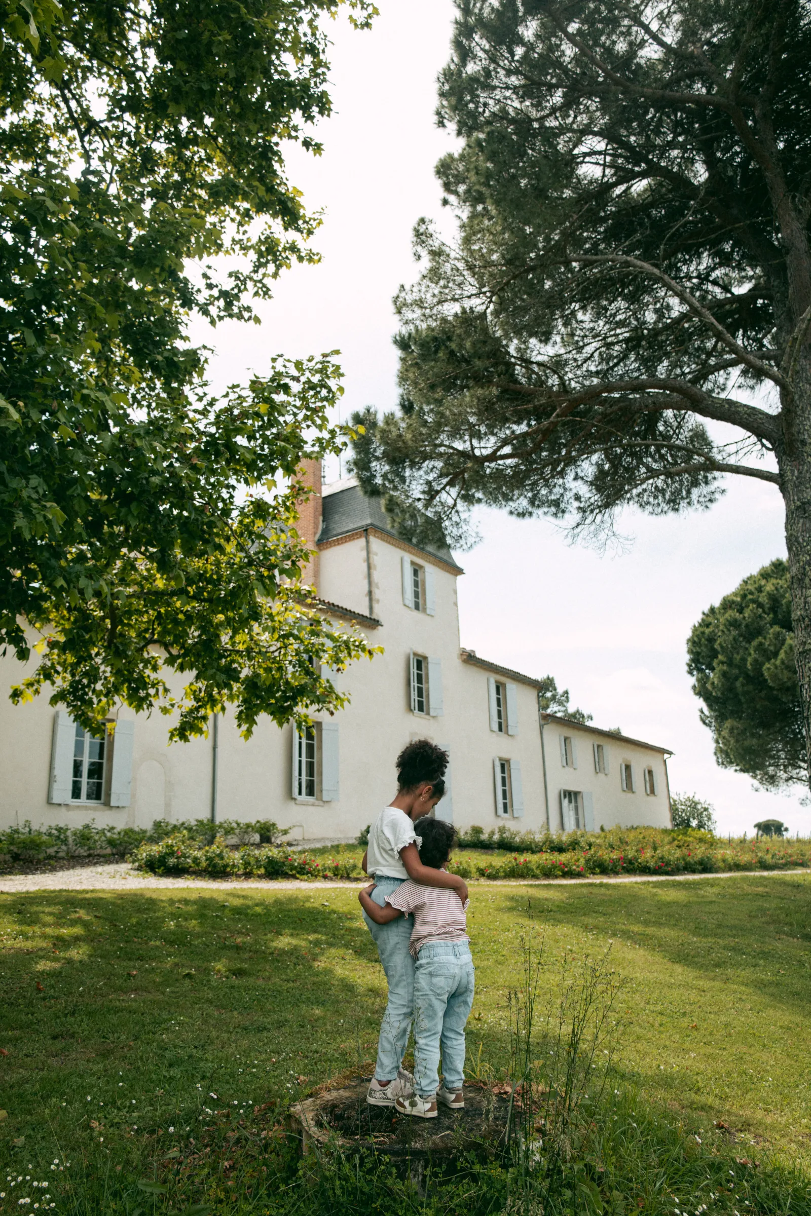 Two children standing beneath a large tree in front of a white historic mansion surrounded by green grounds at Malagar estate