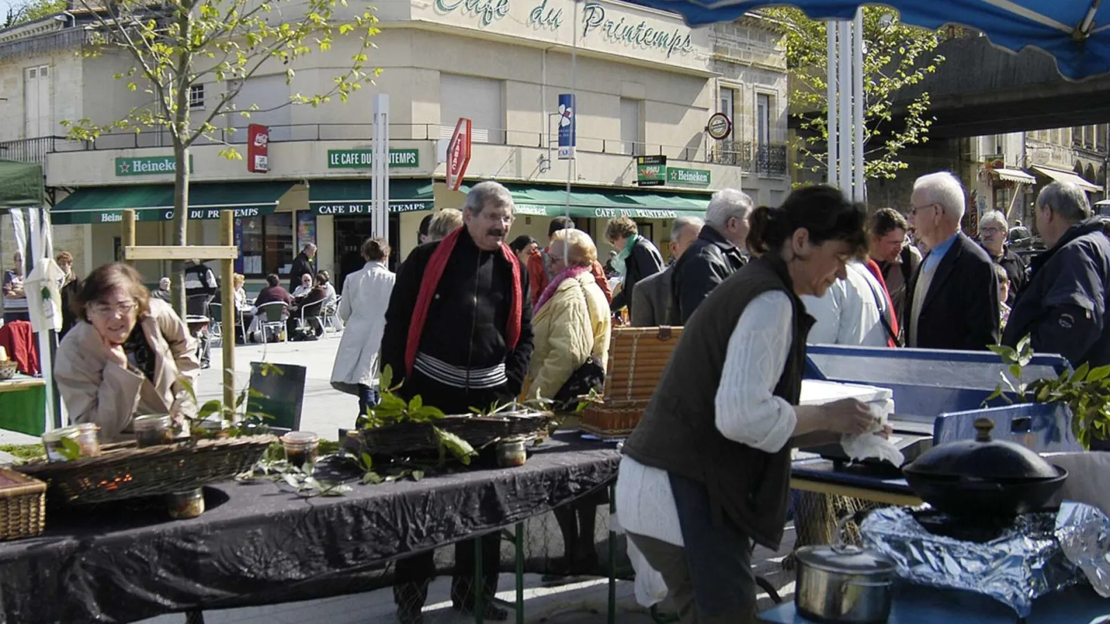 Outdoor market in Lormont with vendors and customers around fresh produce stalls, café and shop facades visible in the background under sunny weather.