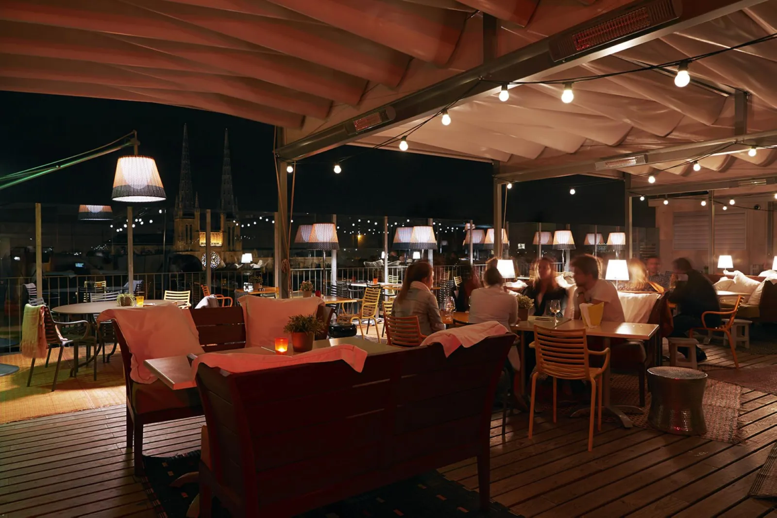 Covered terrace of Mama Shelter Bar in Bordeaux at night, featuring wooden tables and chairs, designer lamps, seated guests, red and grey furnishings, with illuminated cityscape in the background.