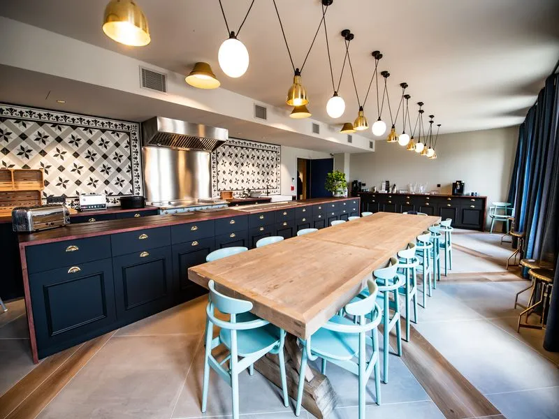 Modern professional kitchen featuring a wooden central island with turquoise blue chairs, navy cabinetry, black and white geometric backsplash, and suspended gold pendant lights.