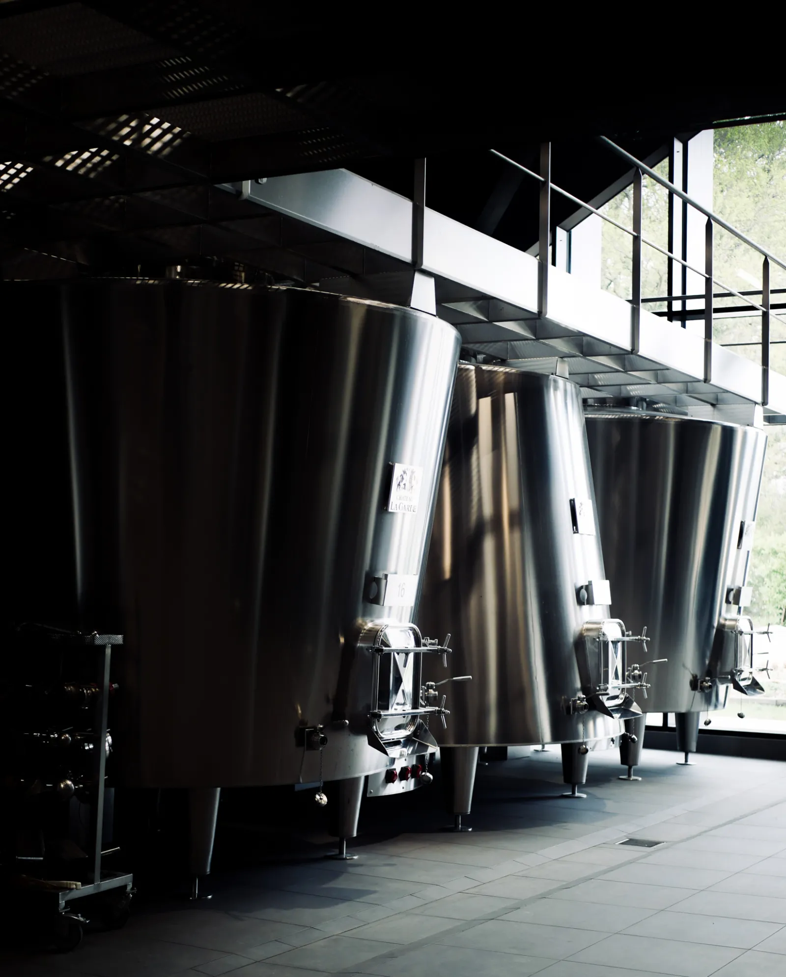 Three stainless steel fermentation tanks lined up in a winery cellar at Château La Garde in Bordeaux, under a covered structure with metal beams and natural light.