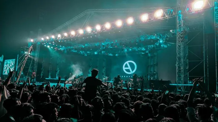 Concert crowd at an electronic music festival in Bordeaux's submarine base, with turquoise blue lighting and metal stage structure.