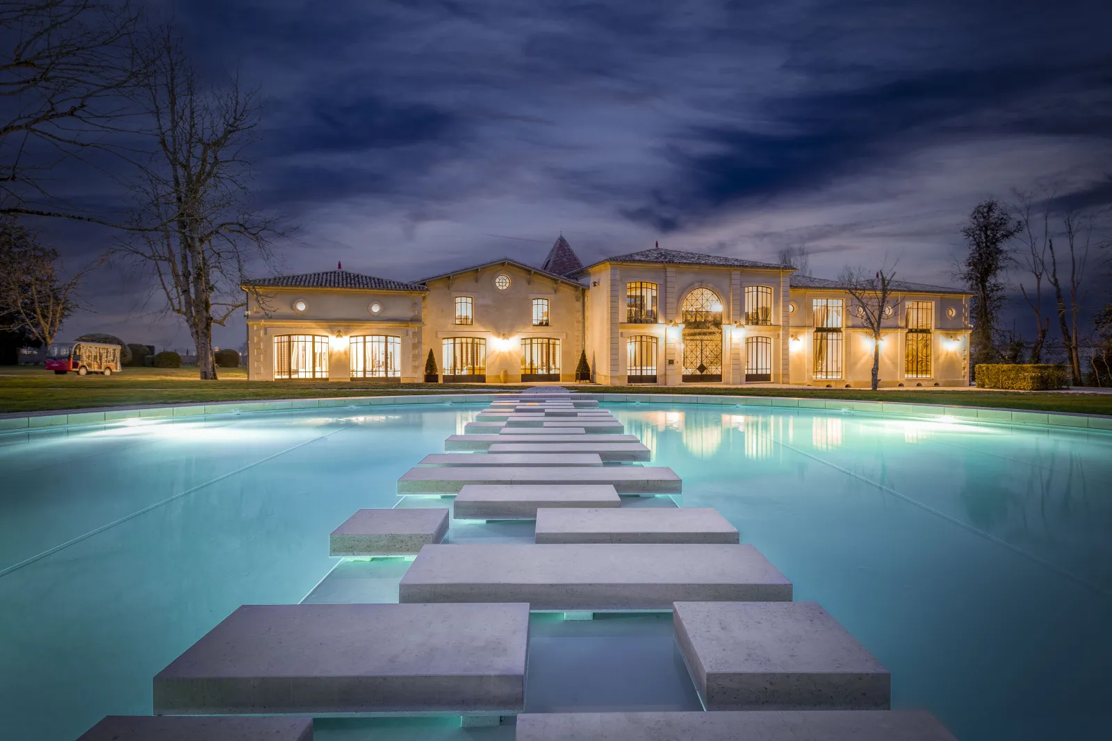 Night view of Château Montlabert in Bordeaux with its golden-lit façade, adjacent to a turquoise pool crossed by a symmetrical gray stone pathway, beneath a dramatic twilight sky.