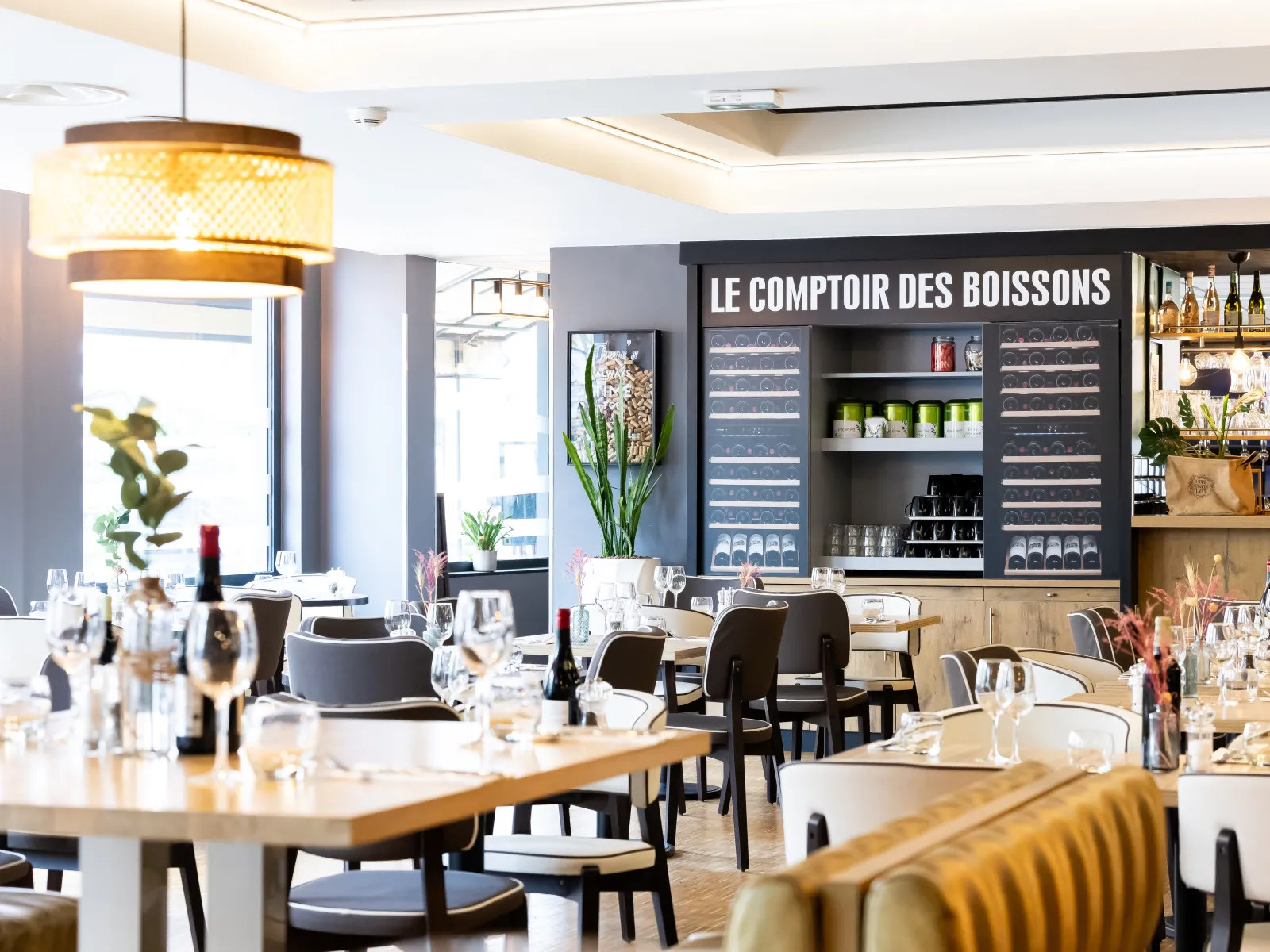 Le Comptoir des Boissons restaurant at Ibis Bordeaux Centre Mériadeck, featuring wooden tables, black chairs, bottle bar, yellow pendant lighting, and large windows.