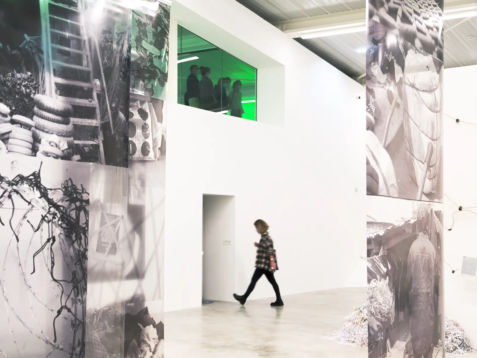 Interior of FRAC Nouvelle-Aquitaine MECA in Bordeaux with a person walking between large black and white photographic panels displayed on white walls, with a green-lit window in the background.