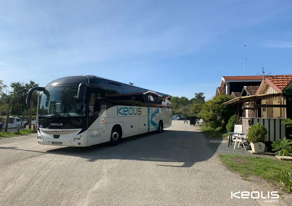 A white and black Keolis coach bus parked in front of a half-timbered building in the Bordeaux region, with blue sky in the background.