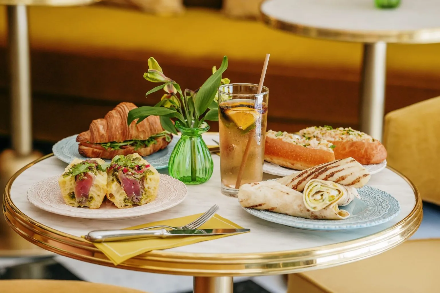 Round table with golden rim displaying an elegant breakfast assortment: croissant, Eggs Benedict with ham, buttered toast, glass of iced lemon water, yellow napkin and green flowers in vase, in a refined café setting in Bordeaux.