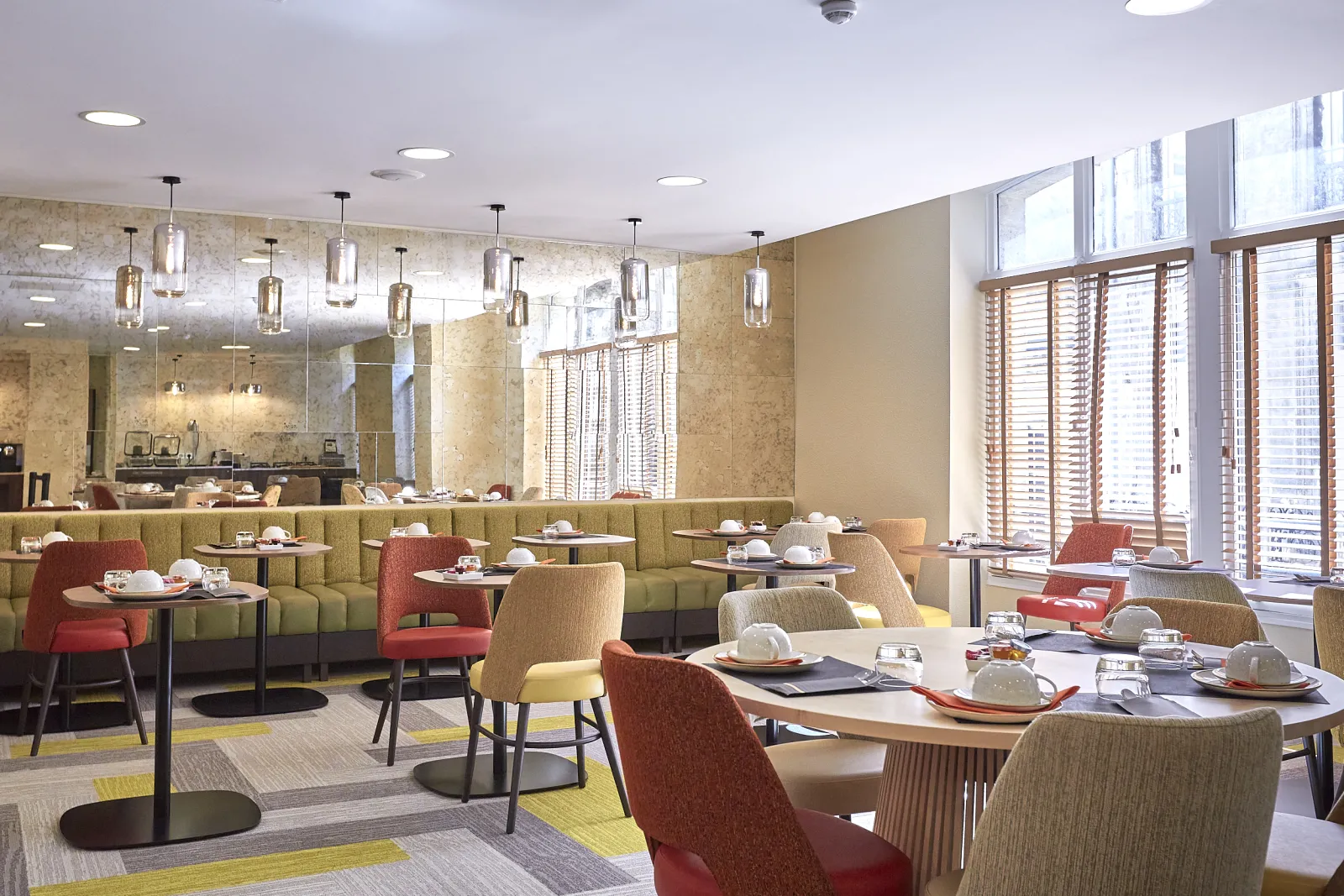 Elegant restaurant at Quality Hôtel Bordeaux Centre featuring green banquettes, red and beige chairs, black tables, modern pendant lights, large windows with wooden shutters, gray and yellow flooring, and a bar counter in the background.