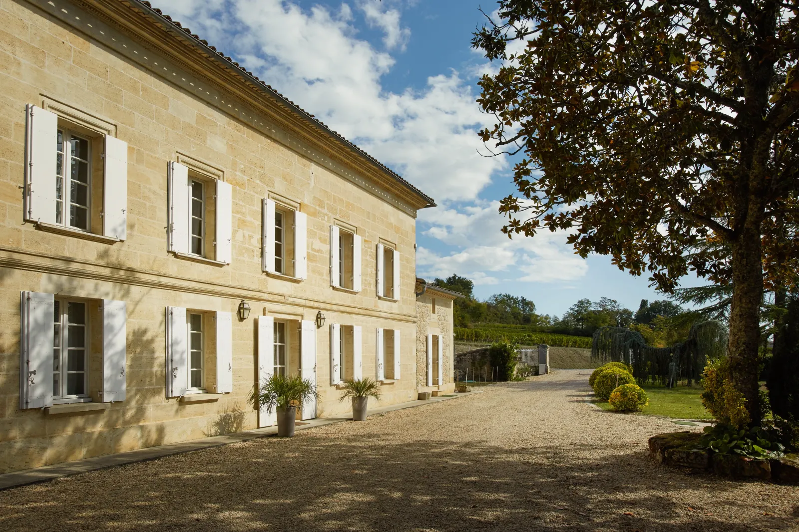 Elegant facade of Château Bernateau in Bordeaux, stone building with white shutters, gravel driveway and vineyard landscape in the background