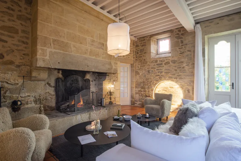 Elegant living room in Château Toulouse Lautrec featuring a stone fireplace with active fire, white sofa, grey armchairs, exposed beam ceiling, modern pendant lights, and glazed doors opening to the garden.