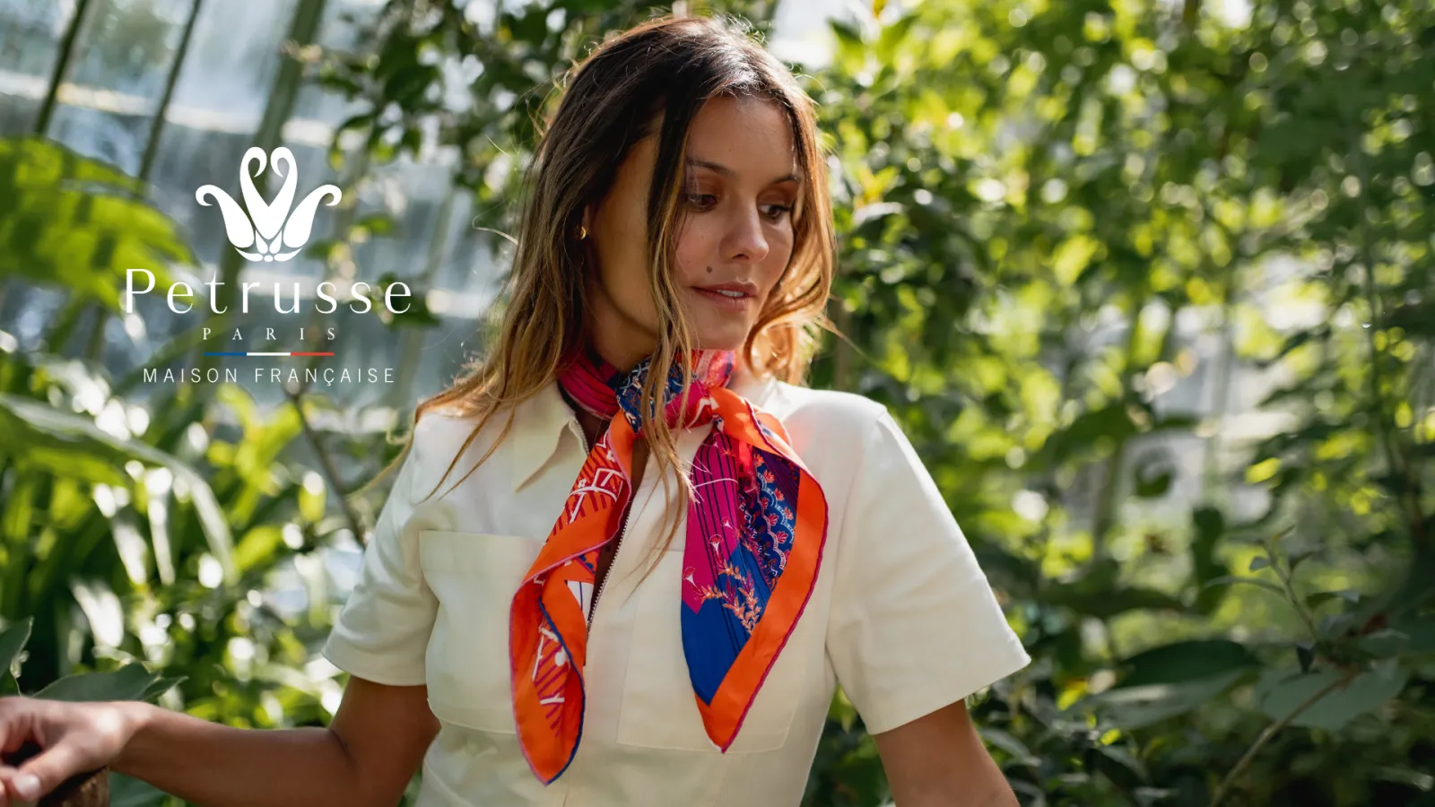 Woman wearing a white short-sleeved shirt with a colorful red and blue scarf, posing in a lush green garden in Bordeaux for Petrusse Maison Française