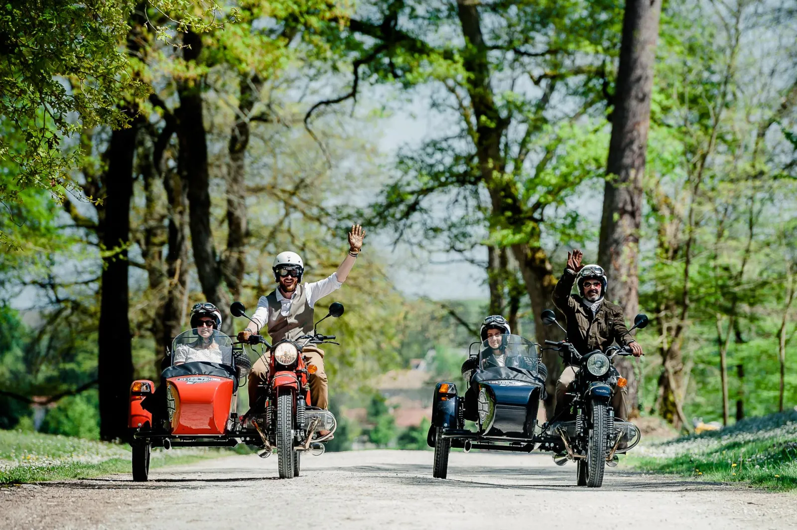 Five riders on vintage motorcycles with sidecars traveling down a tree-lined road, one waving their hand, during a sightseeing tour in Bordeaux.