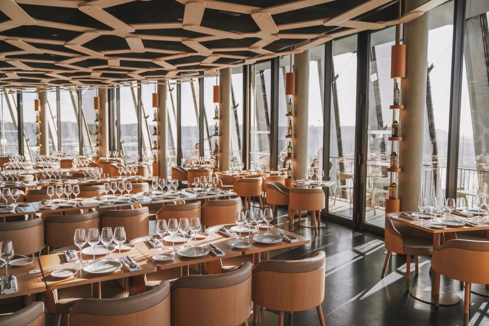 The 7 panoramic restaurant at La Cité du Vin in Bordeaux featuring elegant light wood tables, caramel-colored chairs, wine glasses, modern geometric ceiling, and floor-to-ceiling windows overlooking the city