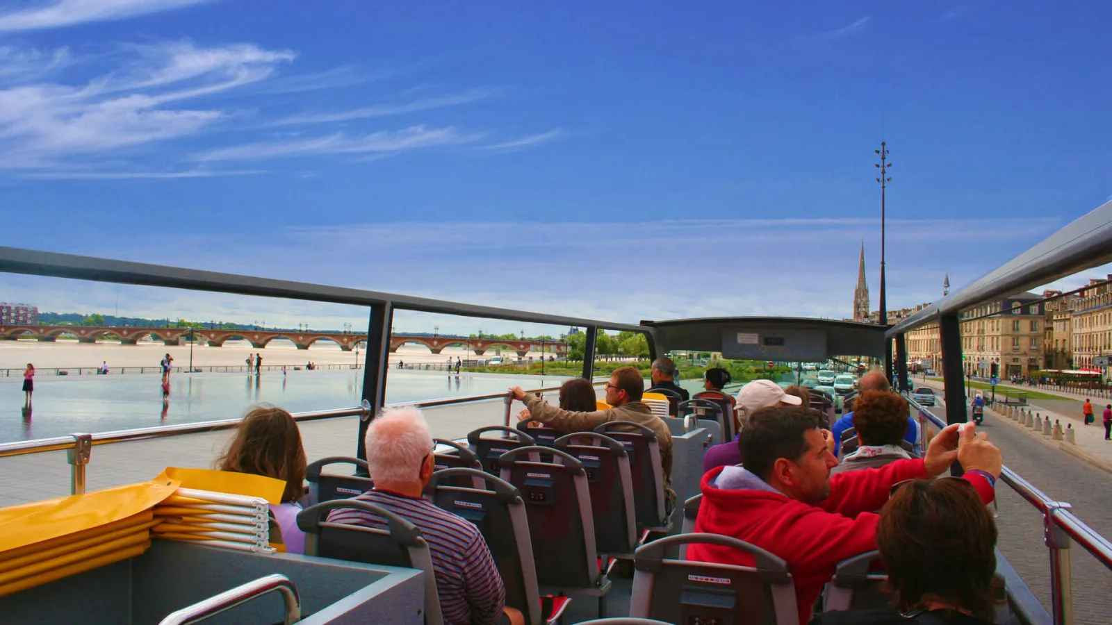 View from an open-top tourist bus of the Bordeaux Visiotour with passengers looking at the Garonne river, bridges, and historic architecture of Bordeaux under a blue sky.