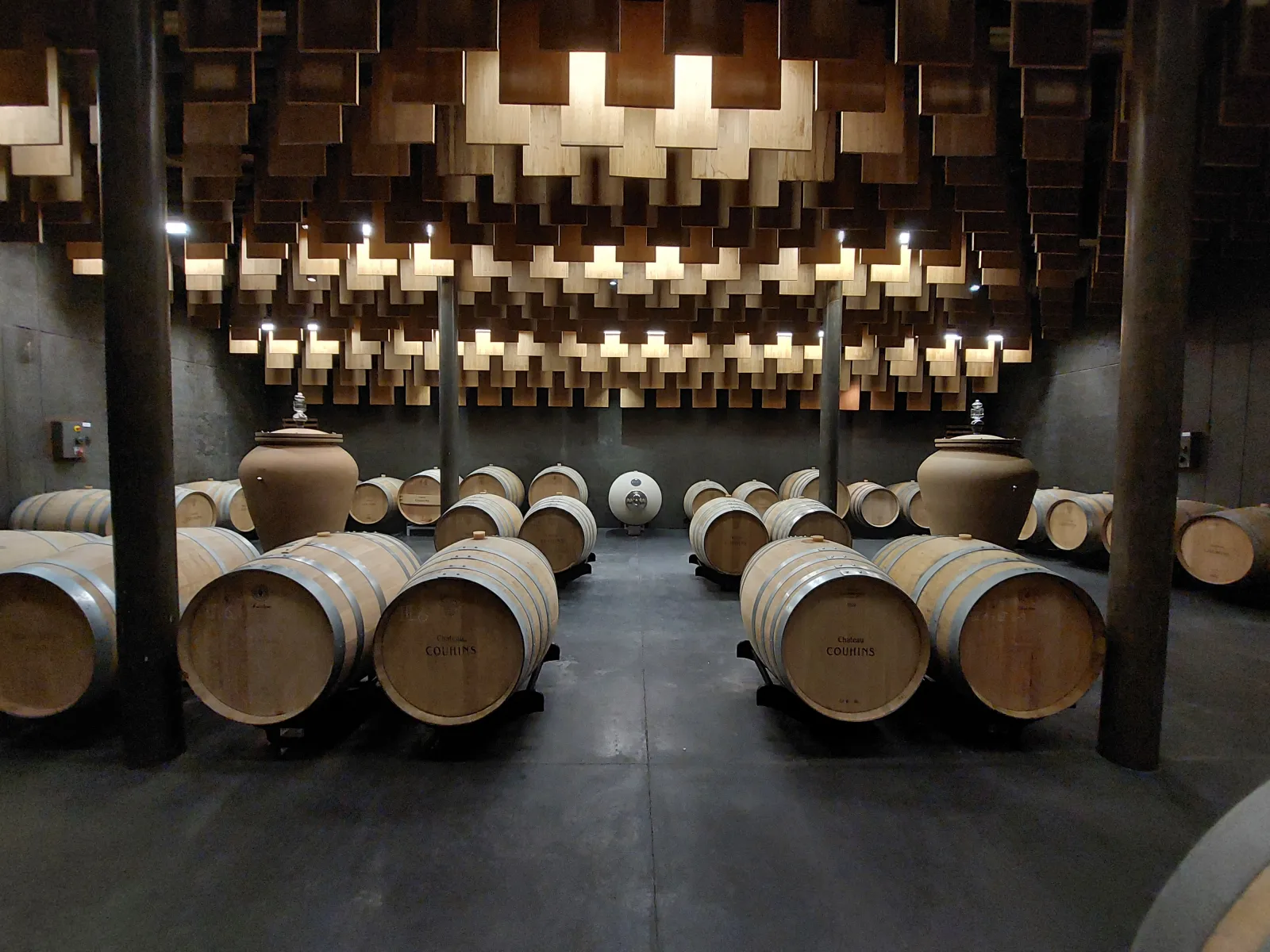 Aging cellar of Château Couhins in Bordeaux featuring oak barrels lined up on the floor and stacked casks in the background under a modern architectural ceiling with wooden panels.