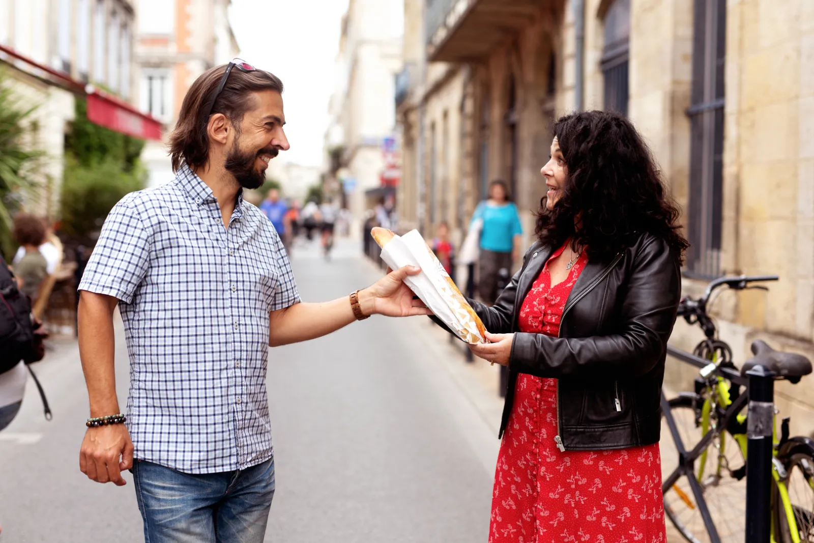 A smiling man and woman exchange a sandwich on a paved street in Bordeaux, with historic buildings and pedestrians in the background.