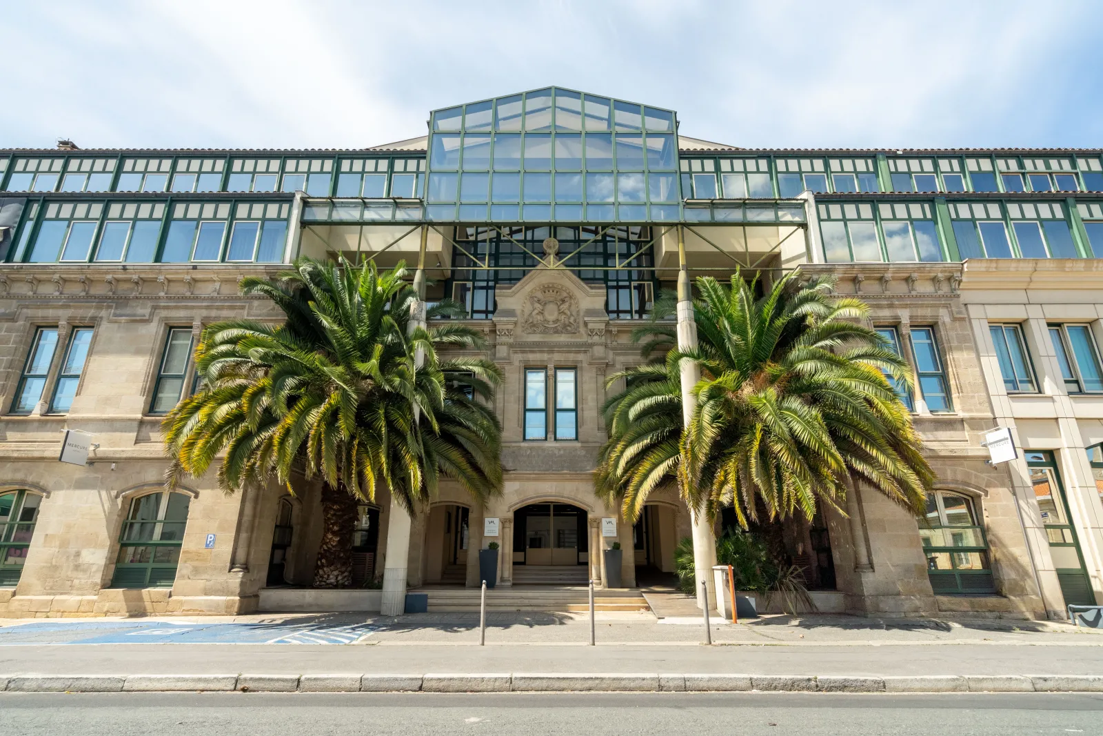 Front façade of Mercure Bordeaux Château Chartrons hotel in Bordeaux, featuring a historic stone building with modern glass roof structure and two palm trees
