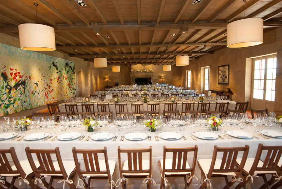 Elegant reception hall at Château Haut-Bailly in Bordeaux featuring white-dressed tables, wooden chairs, exposed beam ceiling, cylindrical pendant lights, and colorful wall artwork.