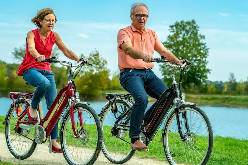 Senior couple riding electric bikes on a path near water in Bordeaux, sunny day with trees in the background