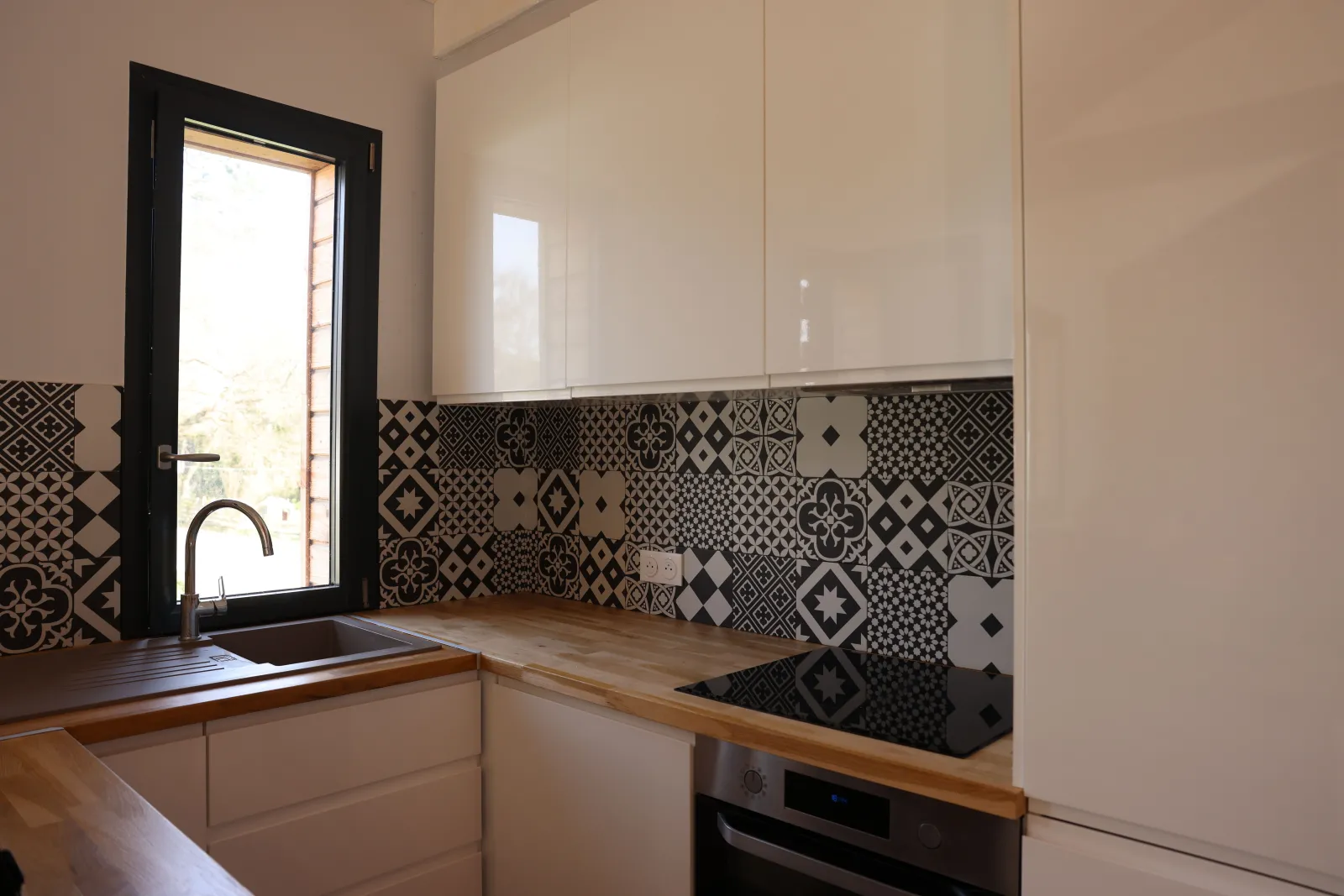 Modern kitchen with white cabinetry, geometric black and white patterned backsplash tiles, light wood countertop, sink under black-framed window, and door opening outside. Éco-Cottage Alfred, Château Loudenne, Bordeaux.