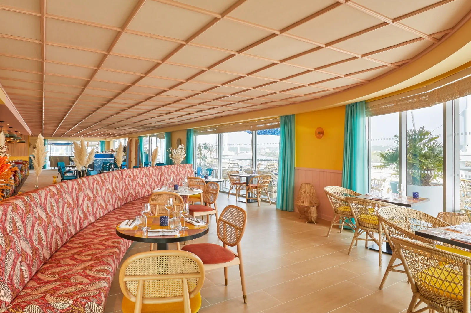 Bright modern lounge at Hôtel Indigo Bordeaux Centre Chartrons featuring rattan furniture, coral pink seating, geometric ceiling design, and floor-to-ceiling windows overlooking Bordeaux.
