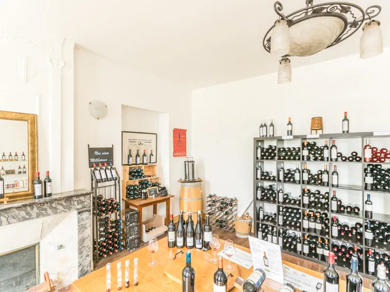 Interior of a wine cellar featuring a marble counter, metal shelves stocked with wine bottles, wooden barrels, tasting glasses, and wine product displays in Bordeaux.