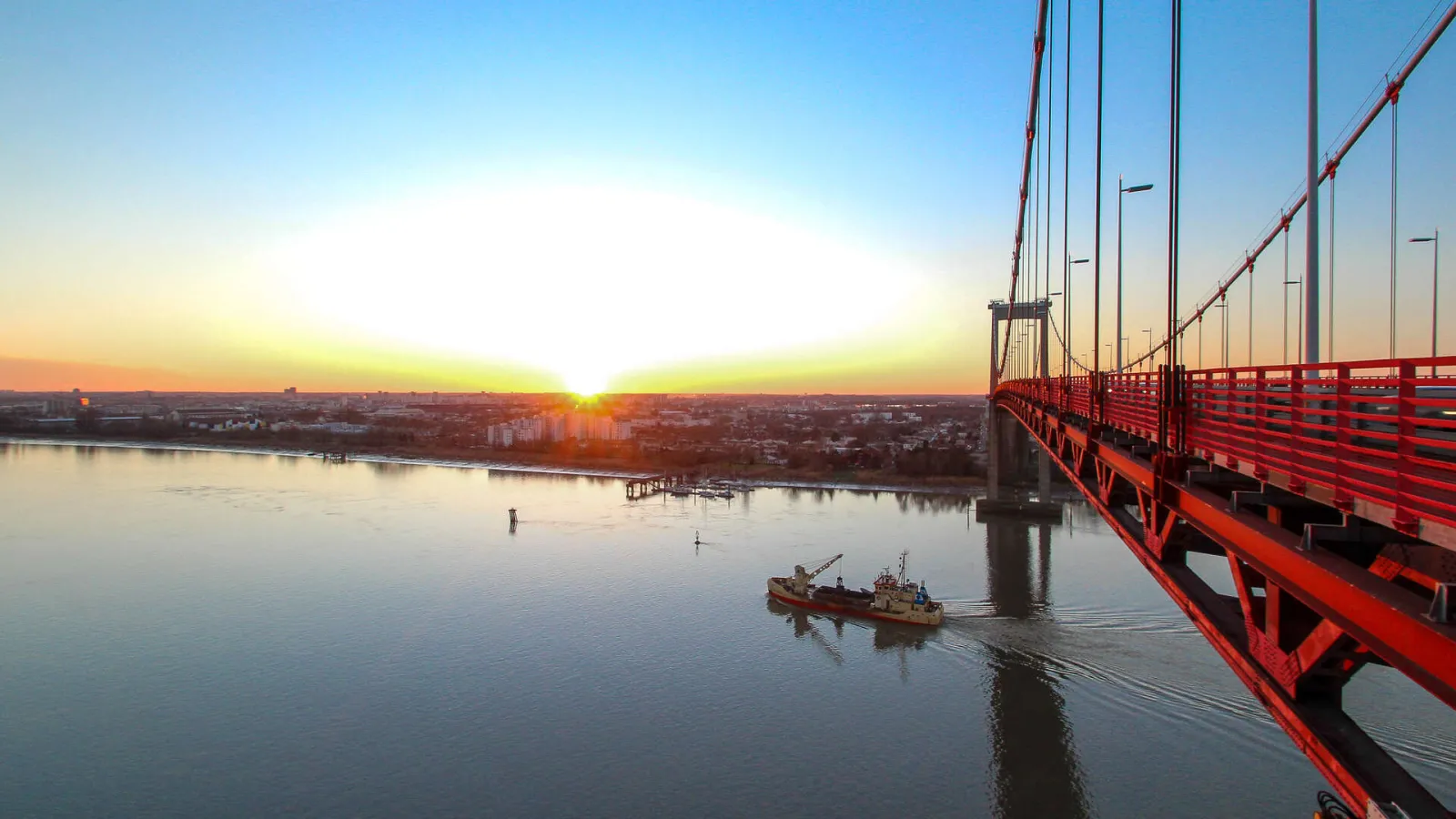 Sunrise view of the Aquitaine Bridge in Bordeaux, with a vessel passing under the red suspension bridge and the city reflected in the calm waters of the Garonne River.