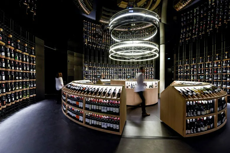 Modern wine cellar interior featuring wall shelving stocked with wine bottles, a central tasting counter, a contemporary ring-shaped light fixture, and two customers browsing the selection in Bordeaux.