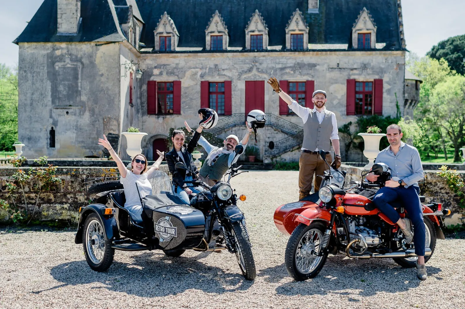 Six people cheerfully posing in front of a historic castle with vintage motorcycles and sidecars, wearing helmets and raising their arms in celebration during a retro tour.