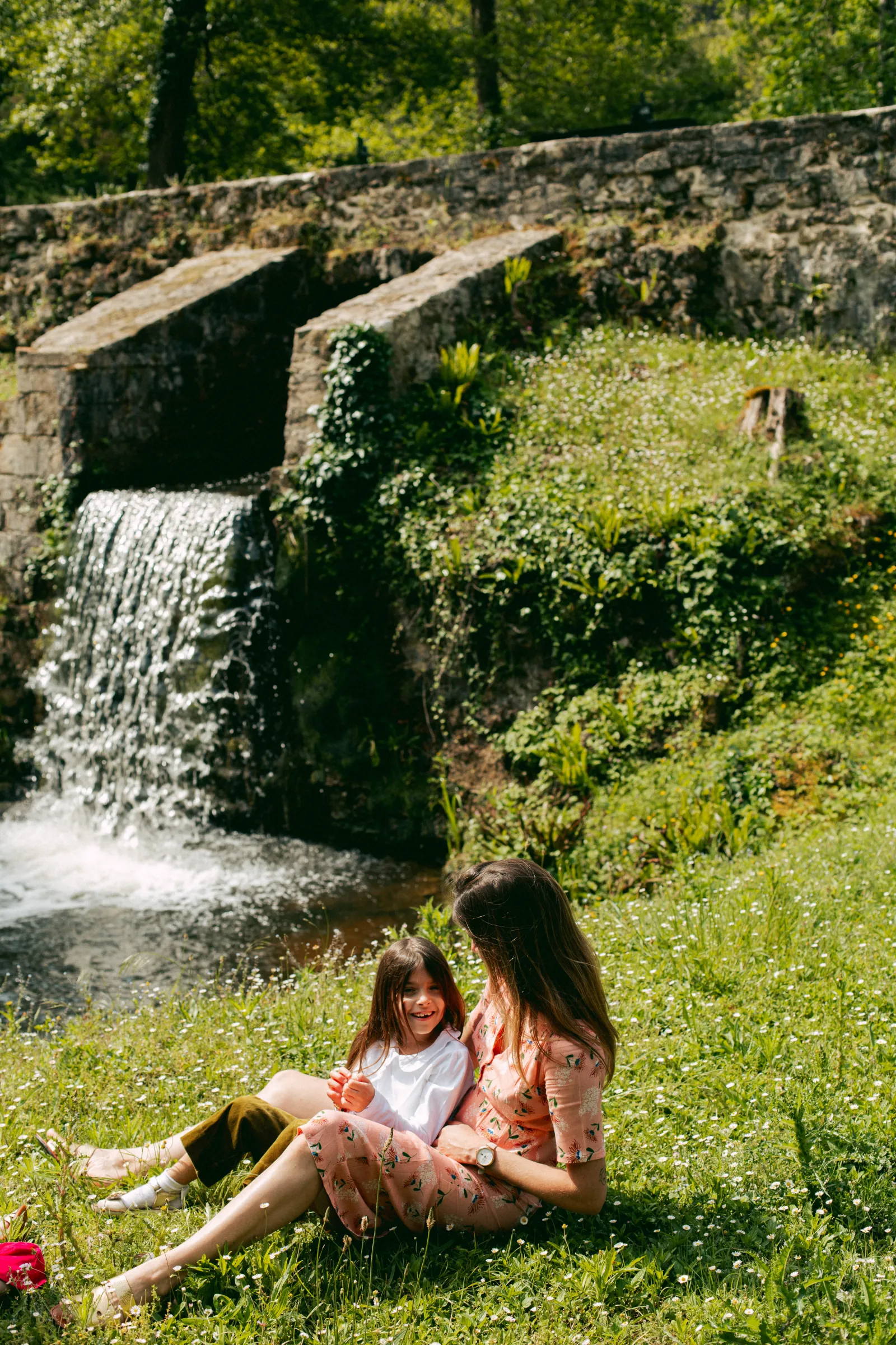 Two children sitting on grass in front of a waterfall and ivy-covered stone wall at the Royal Castle of Cazeneuve near Bordeaux.