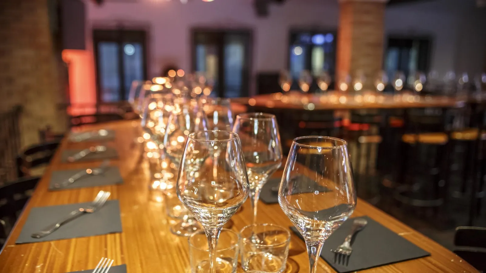 Elegant restaurant table with wine glasses in a row, cutlery, grey napkins and lit candles, in a modern dining room with pink walls and brick details.