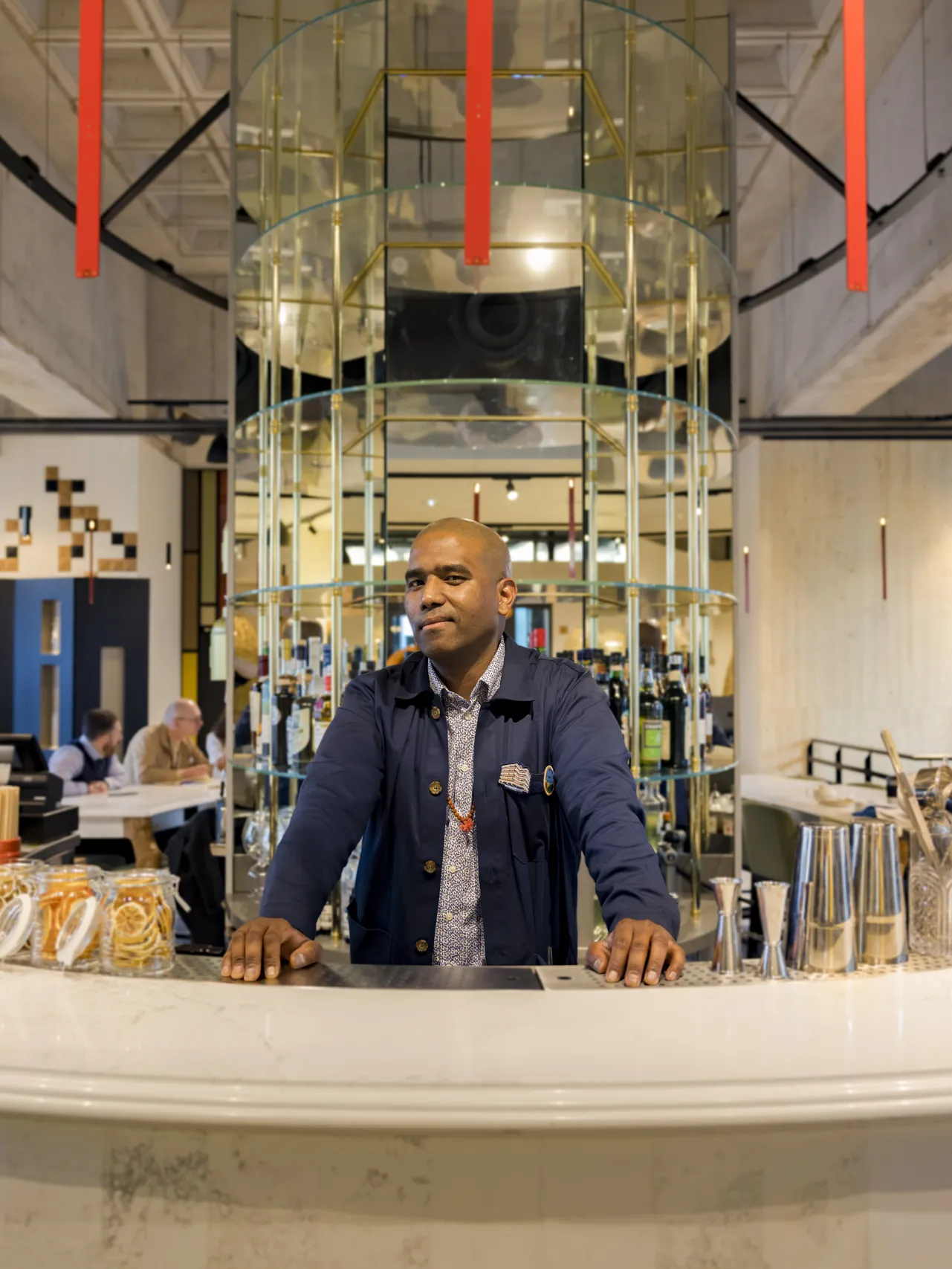 A man wearing a dark blue jacket stands at a white counter inside the BADA in Bordeaux, a modern space featuring a glass structure and red columns in the background.