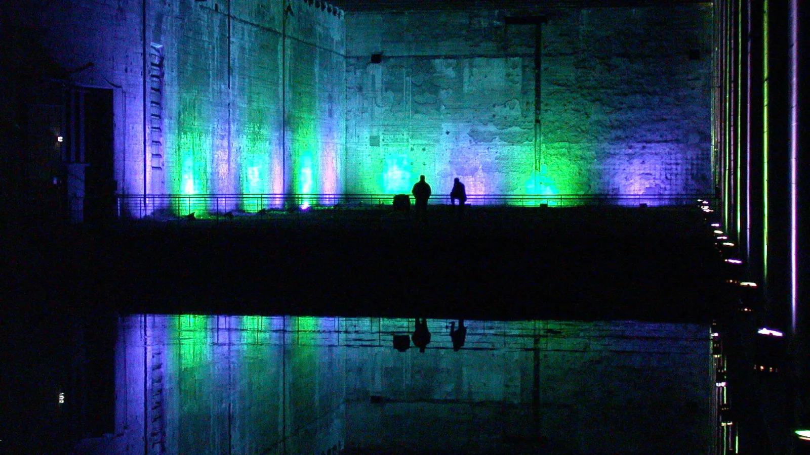 Bordeaux's submarine base lit up at night with blue, green, and purple spotlights, reflecting on the water. Two people silhouetted on a platform.