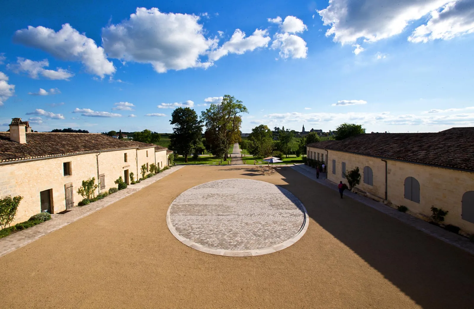 Inner courtyard of Château Soutard featuring stone buildings, circular paving pattern, and tree-lined pathway beneath clear blue sky