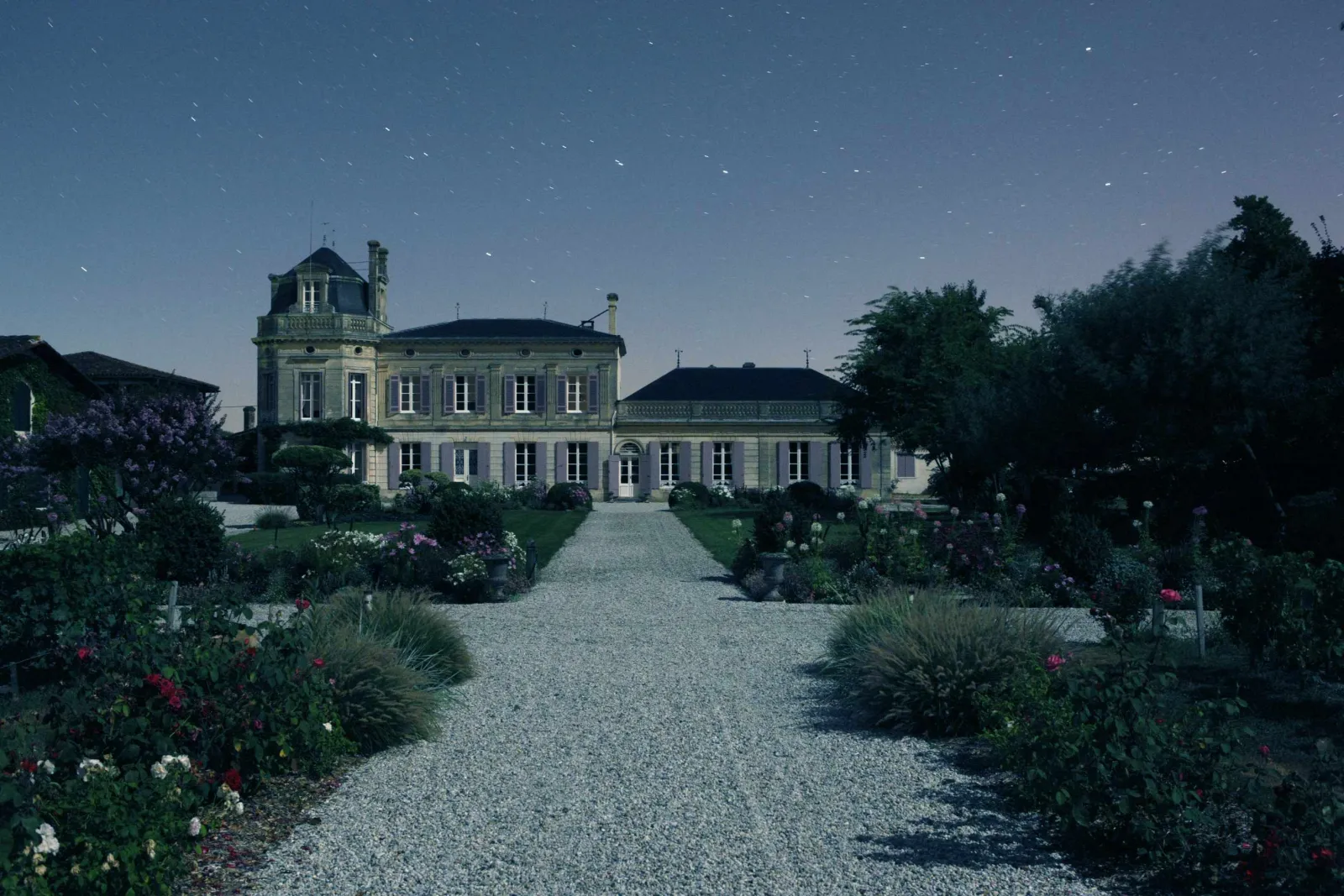 Night view of Château Chasse-Spleen in Bordeaux, an elegant manor house with white facades and a dome, surrounded by flowering gardens and a gravel pathway under a starlit sky.