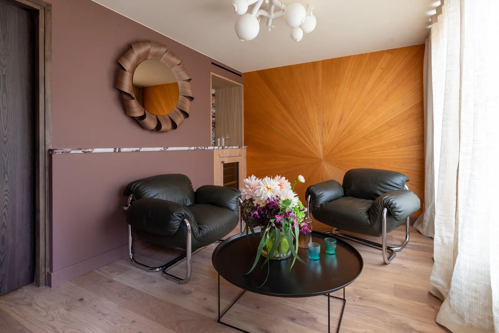 Modern lounge at Yndo Hotel in Bordeaux featuring two black leather armchairs, a round black coffee table, mauve-pink and orange wood walls, a circular mirror and fresh flowers.