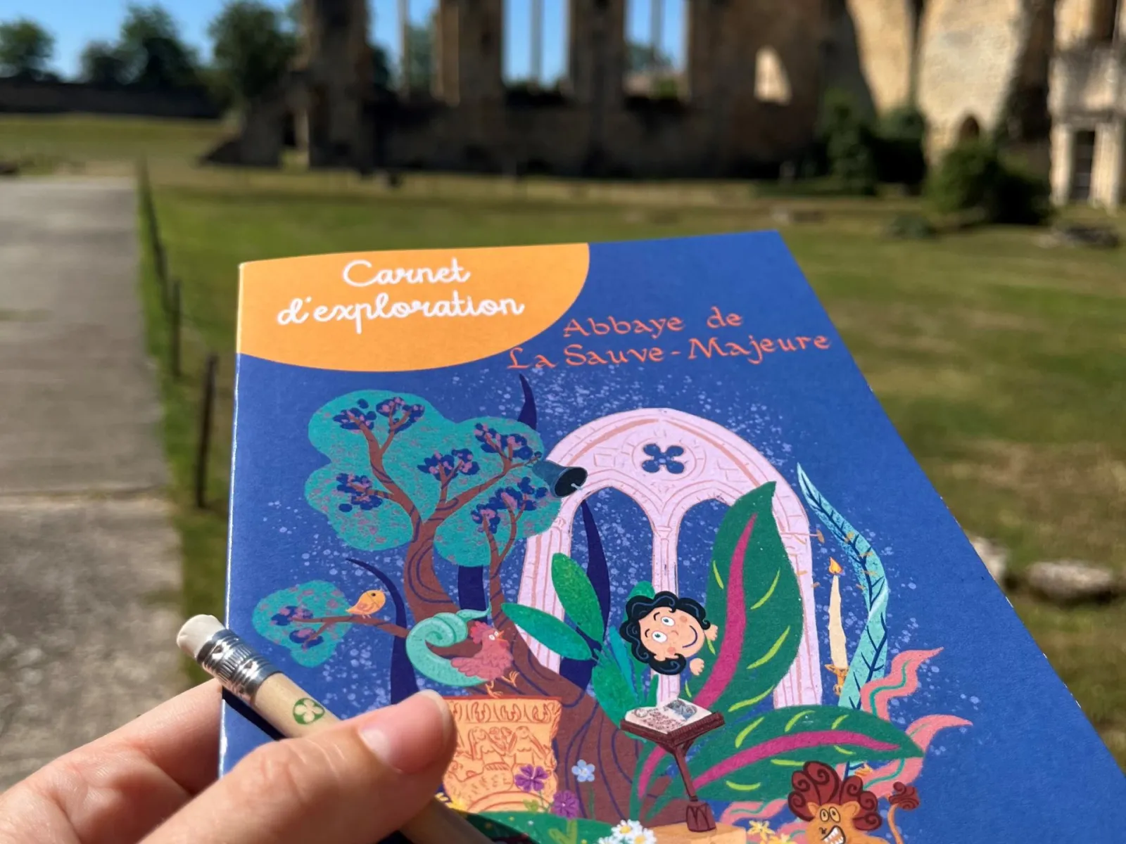Exploration notebook for the Abbey of La Sauve-Majeure held in hand in front of the abbey ruins in Bordeaux, featuring colorful artwork of a child exploring a fantastical landscape.