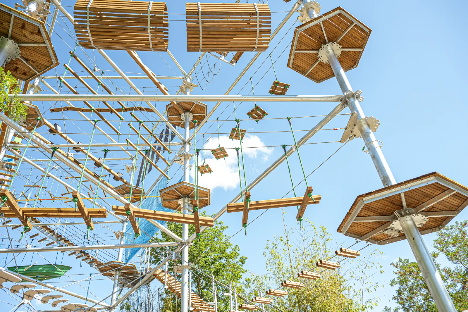 Overhead view of a wooden adventure park structure featuring suspended courses, metal beams, and aerial game elements interconnected with ropes and cables against a clear blue sky.