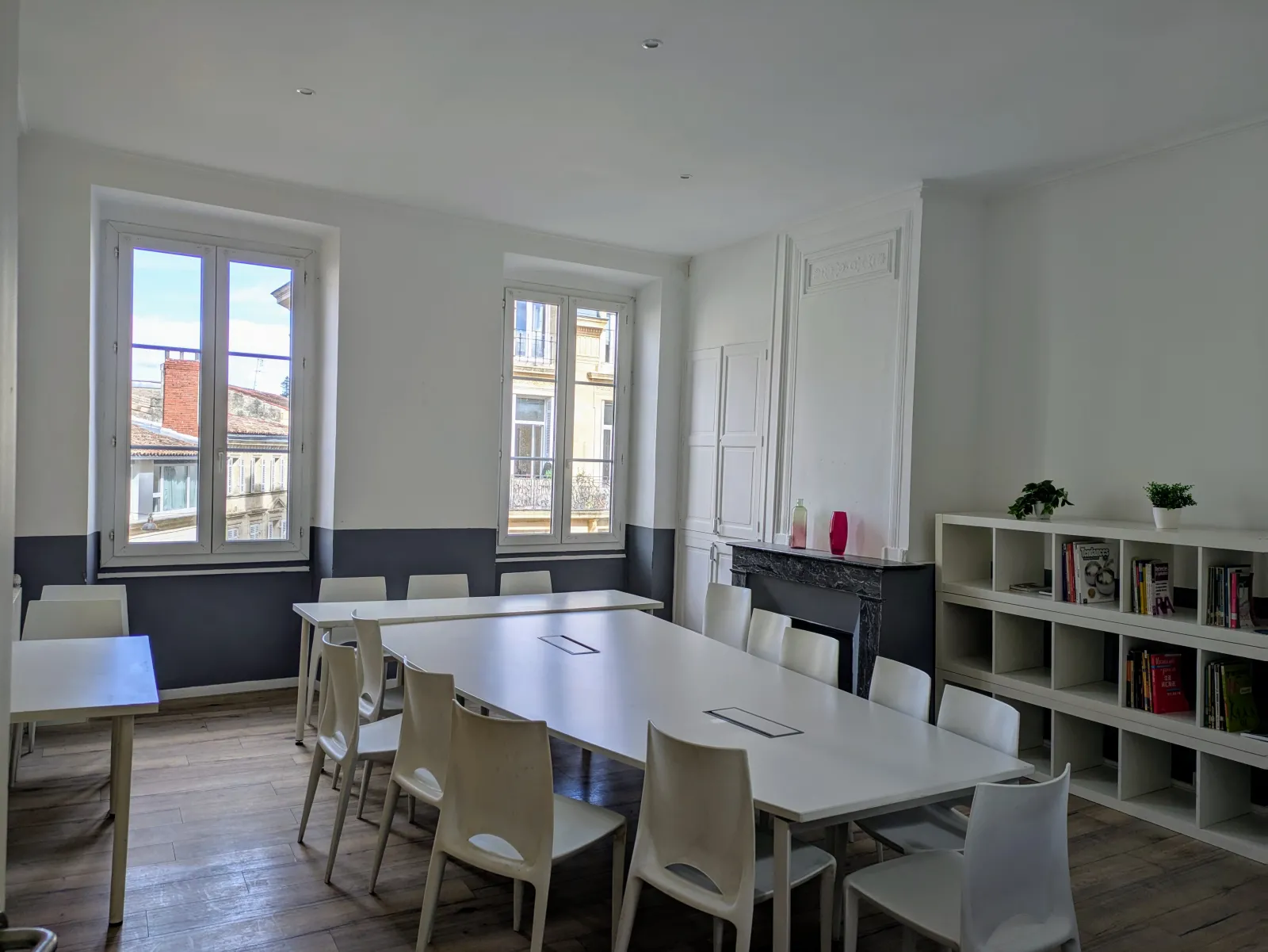 Modern workspace at FL Studios in Bordeaux featuring white tables, beige chairs, built-in shelving, black fireplace, and large windows overlooking the city.