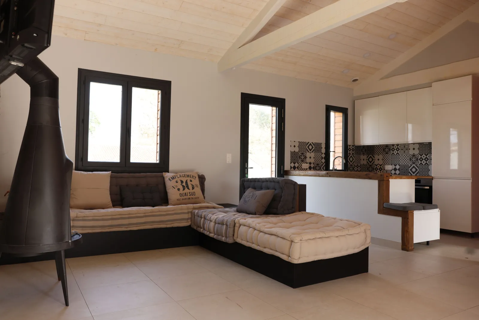 Modern interior of an eco-cottage featuring an open-plan living area, fitted kitchen, exposed beam ceiling, black-framed windows, beige and black furnishings, and a cylindrical black stove.