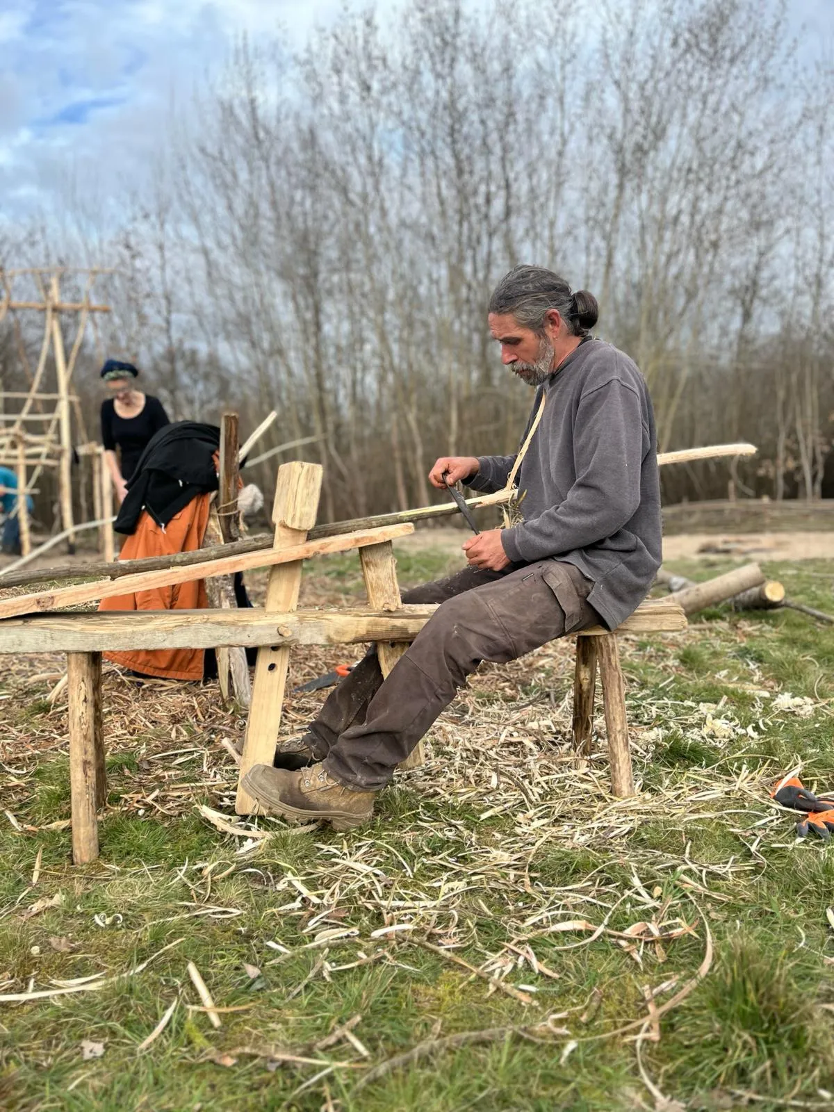 Two craftspeople work with wood at a workbench during a medieval historical reenactment. A man in the foreground shapes a wooden piece with a tool while a woman observes in the background. Bare poplar trees line the reconstructed medieval site.