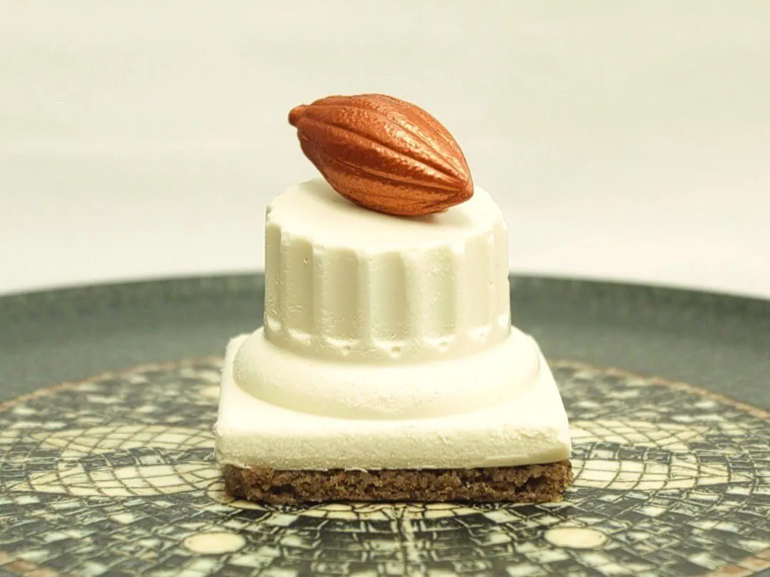 Square cake with a biscuit base, topped with white cream and a roasted almond, served on a gray plate