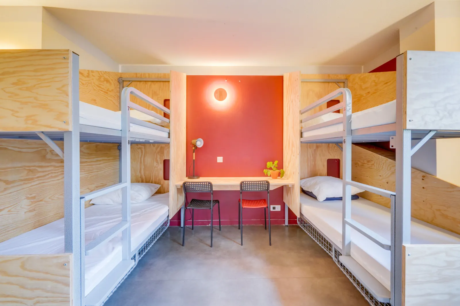 Hostel dormitory room in Bordeaux featuring four wooden and metal bunk beds, red and beige walls, and a central work area with two chairs and a desk