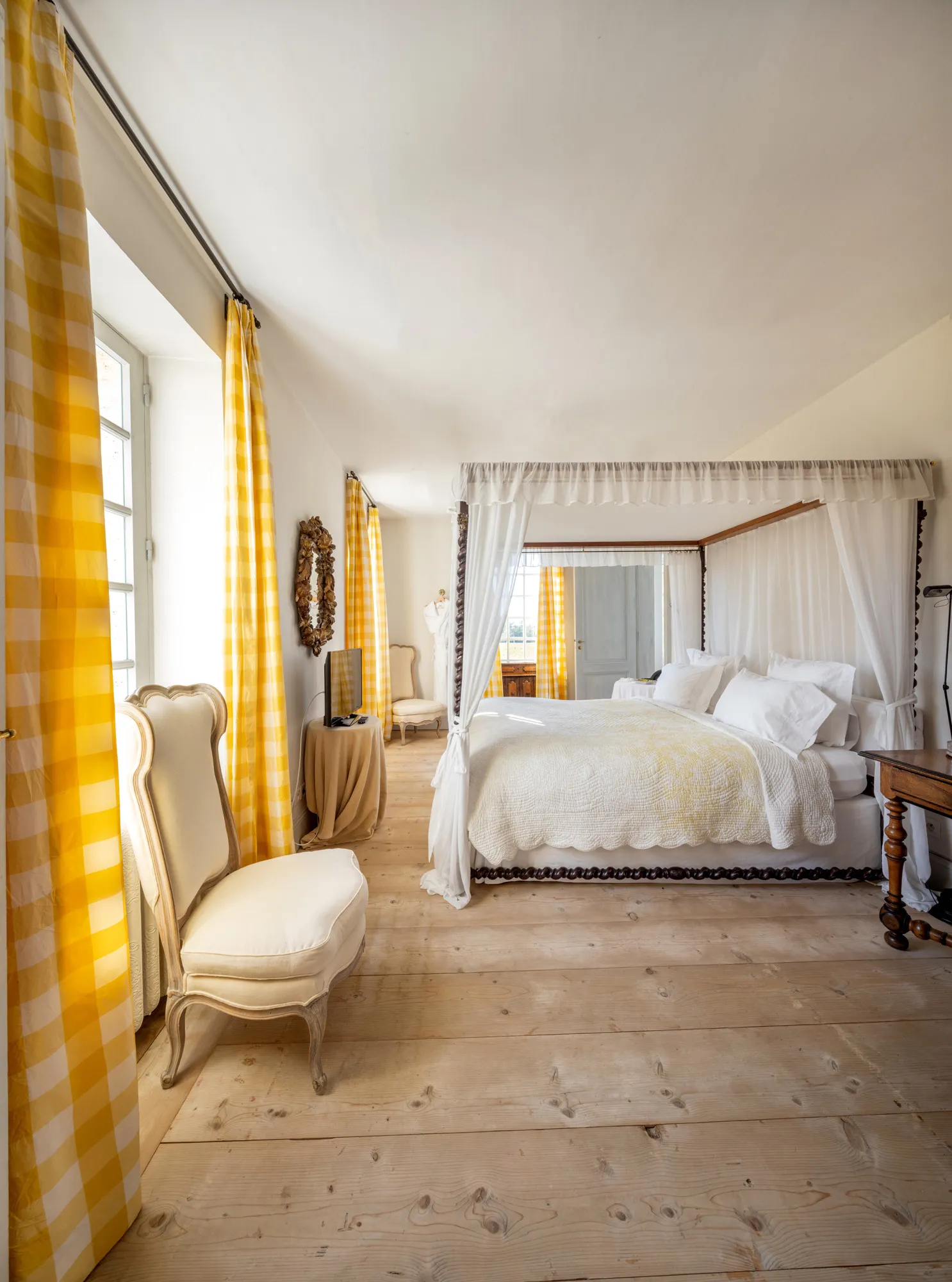 Elegant guest room at Château du Tertre in Bordeaux, featuring a white canopy bed, yellow checked curtains, cream-colored armchair, light wooden floors, and refined decor.