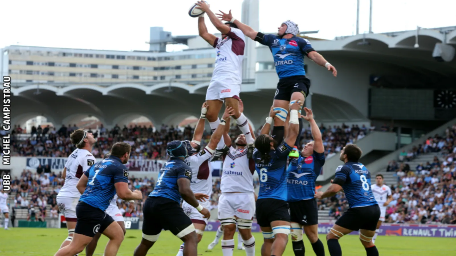 Rugby match in Bordeaux: two players jump for the ball, one in white and maroon jersey, the other in blue, in front of spectators at Chaban-Delmas stadium.