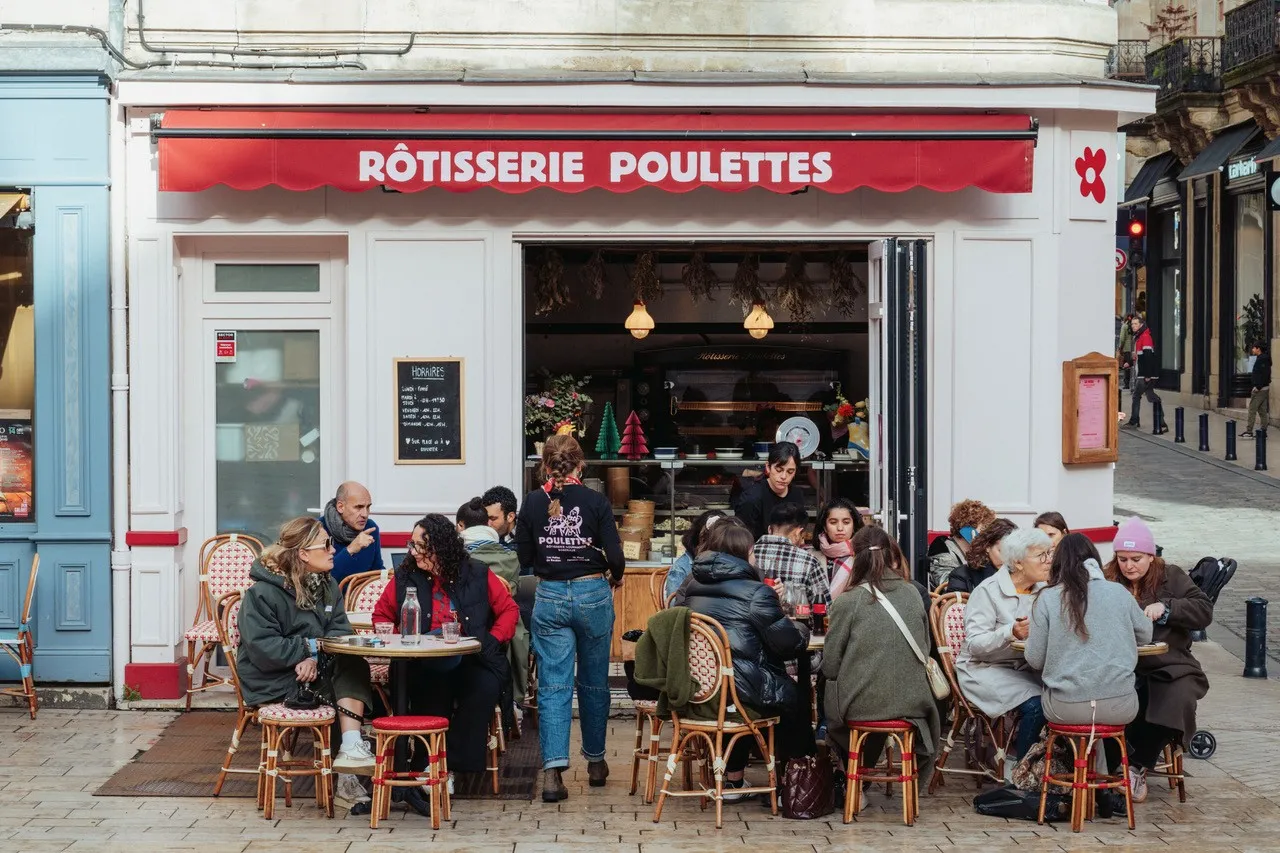 Exterior view of Rôtisserie Poulettes in Bordeaux with a bustling outdoor terrace featuring customers seated at bistro tables enjoying meals in front of the red and white striped storefront.